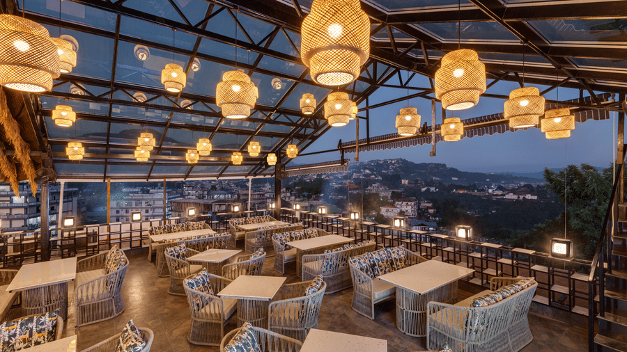An outdoor rooftop restaurant with a large metal and glass structure overhead, woven seating and tables, spherical pendant lights, and a panoramic view of the lights of the town below | Voyage Eastin Suites