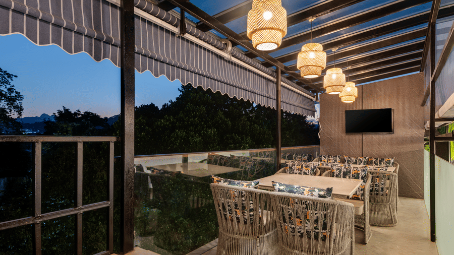 An outdoor rooftop restaurant with a covered patio area and woven seating and tables, spherical pendant lights, and a view of trees under a twilight sky | Voyage Eastin Suites