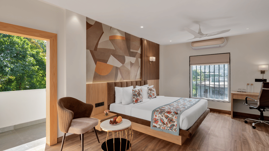 A hotel bedroom with a large bed, light wood flooring, a television, a desk on the right, and a glass door opening to a small balcony with a view of greenery | Voyage Eastin Suites