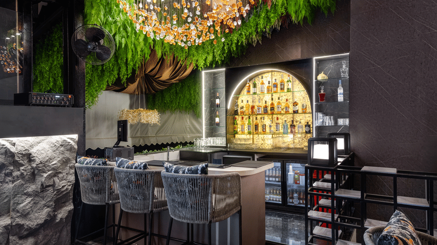 A dark, modern bar area with a lit counter, bar stools, bottles on shelving, and green foliage decorating the ceiling | Voyage Eastin Suites
