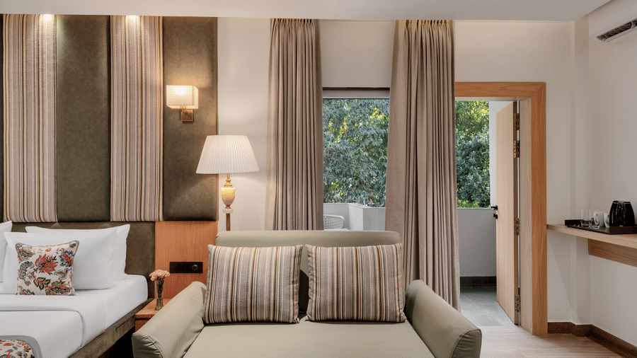 A hotel suite with a sofa seating area near the glass balcony doors, a large bed with floral bedding, and dark wood panelling and curtains | Voyage Eastin Suites