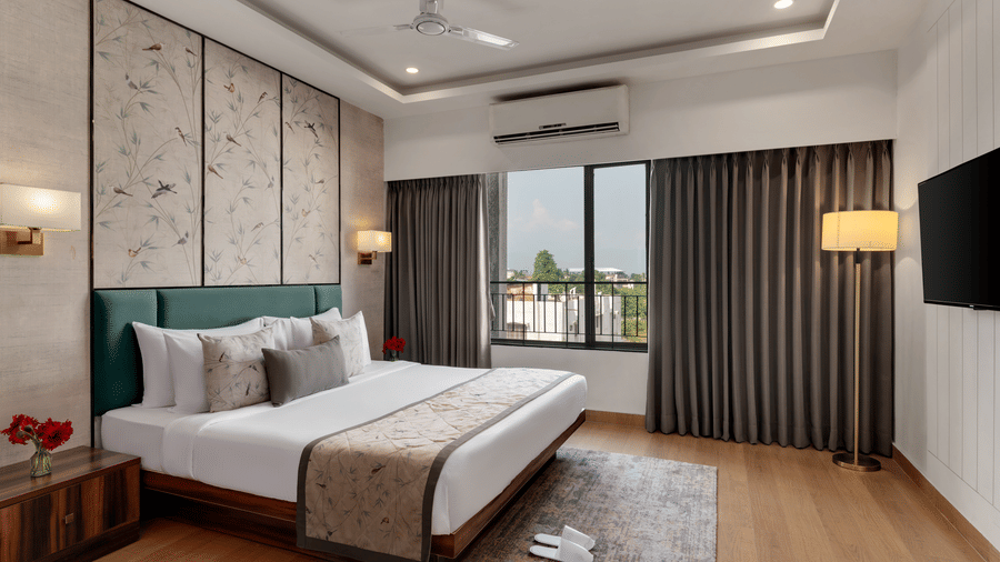 Suite at Voyage Milestone Hotel and Banquet featuring a plush bed by a large window facing the TV on the wall and a floor lamp by the curtains