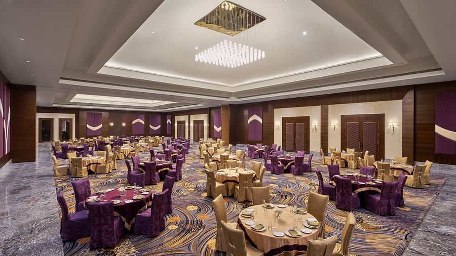Elegant banquet hall featuring a high ceiling with a modern chandelier, richly patterned carpet, and round tables adorned with gold and purple covers - Voyage The Solitaire Bliss.