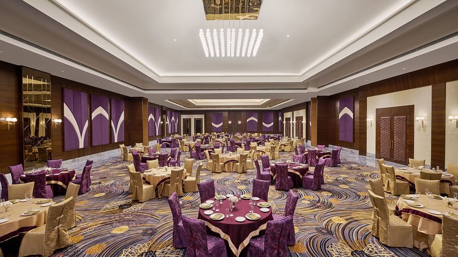 A spacious, elegant banquet hall with round gold-draped tables, purple and gold chairs, and refined place settings. The high ceiling features recessed lighting, a modern chandelier, and purple-gold accents with wood panelling - Voyage The Solitaire Bliss.
