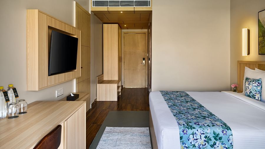 A modern hotel room features a king size bed with white linens and a blue floral runner, a wooden desk with a mounted TV, and an open doorway leading to a closet and bathroom area - Voyage The Solitaire Bliss.