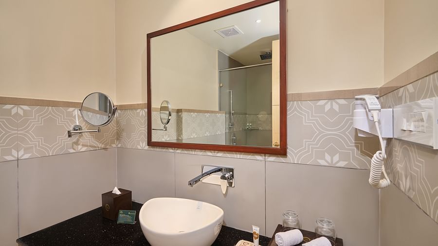 A bathroom with a wood-framed mirror above a white sink on a black counter. Patterned tiles accent the walls. Toiletries, a tissue box, and a wall-mounted hairdryer are visible. A smaller mirror extends from the left wall; the reflection shows a shower - V