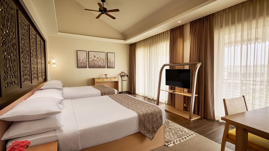 Spacious hotel room with two double beds, a ceiling fan, flat-screen TV, wooden furniture, and large windows with sheer curtains letting in natural light - Voyage The Solitaire Bliss.