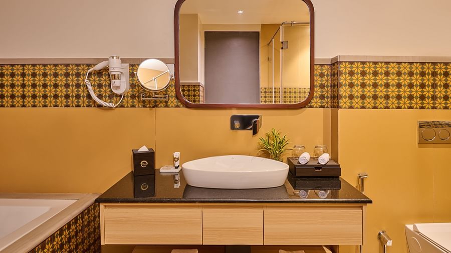 Modern bathroom vanity with a stylish sink, round mirror, wall-mounted hair dryer, and geometric-patterned yellow and brown wall tiles. Neatly rolled towels are placed on the open shelf below the counter - Voyage The Solitaire Bliss.