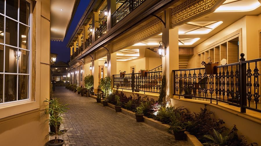 Elegant outdoor corridor at night with warm lighting, potted plants lining a paved walkway, and a balcony-style upper level featuring decorative iron railings and cosy seating areas - Voyage The Solitaire Bliss.
