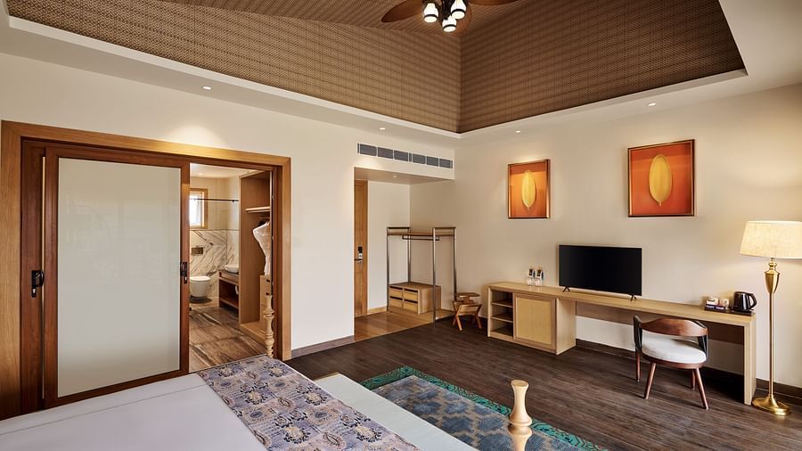 Elegant hotel room featuring a large bed with patterned bedding, wooden flooring, a ceiling fan, wall-mounted TV on a long desk, two framed artworks, and a sliding door leading to a modern bathroom - Voyage The Solitaire Bliss.