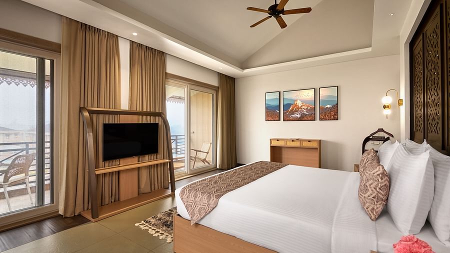 Bright and airy hotel room with a neatly made double bed, wall-mounted TV on a wooden stand, ceiling fan, balcony with outdoor seating, large windows with curtains, and framed artwork above a chest of drawers - Voyage The Solitaire Bliss.