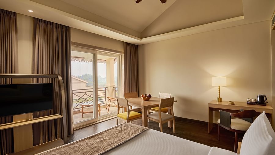 Cosy hotel room with a neatly made bed, flat-screen TV, wooden desk with a lamp and kettle, small dining table with chairs, floor-to-ceiling curtains, and a balcony with a scenic view - Voyage The Solitaire Bliss.