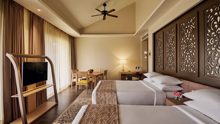 Elegant hotel room with two double beds, carved wooden headboards, ceiling fan, flat-screen TV on a wooden stand, and a seating area near large windows with sheer curtains - Voyage The Solitaire Bliss.