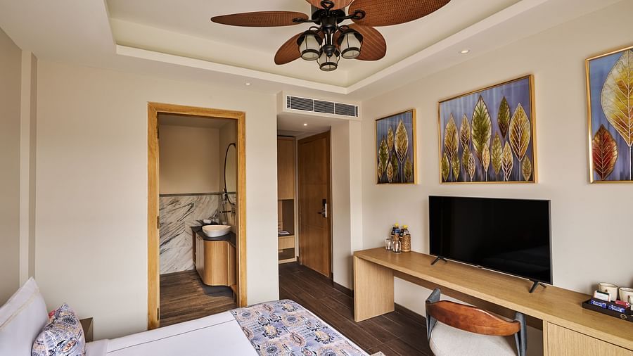 Charming hotel room with a bed, floral bedding, ceiling fan with leaf-shaped blades, wall-mounted TV on a long wooden desk, modern artwork on the wall, and a view into a sleek bathroom with marble accents - Voyage The Solitaire Bliss.