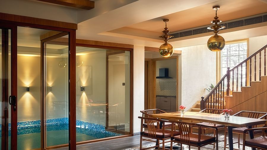 Elegant indoor dining area with a wooden table and chairs, staircase to the side, and a glass-enclosed private pool illuminated with soft lighting - Voyage The Solitaire Bliss.