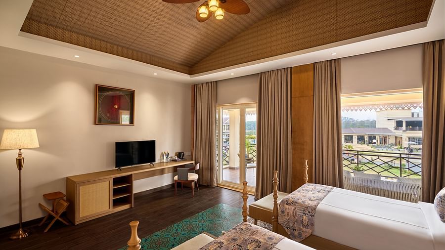 Spacious hotel room with two single beds, wooden ceiling with a fan, a flat-screen TV on a desk, floor lamp, and large windows with curtains overlooking a balcony and garden area outside - Voyage The Solitaire Bliss.
