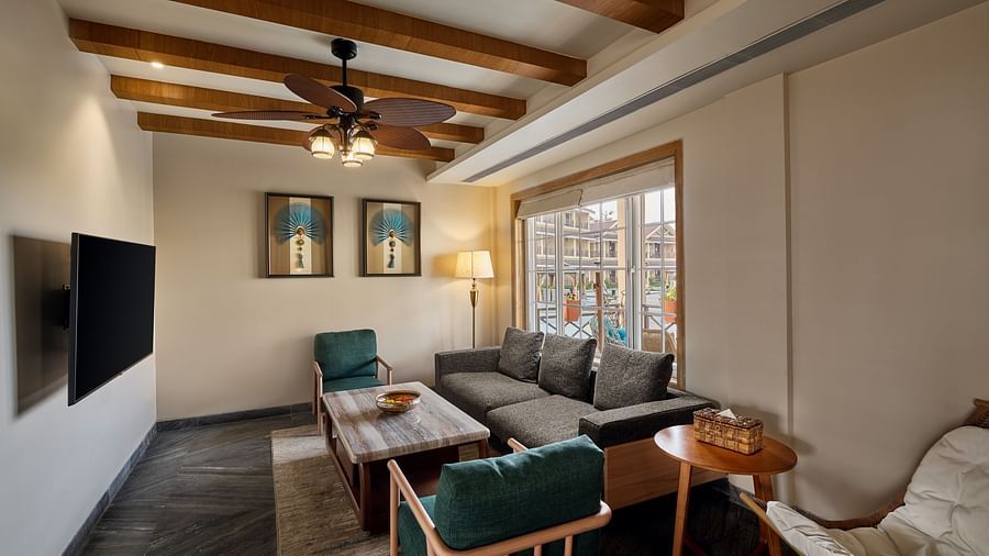 Cosy living room with wooden ceiling beams, a ceiling fan with lights, a grey sectional sofa, green armchairs, a wooden coffee table, and large window allowing natural light. Wall-mounted TV on the left and decorative paintings above the sofa - Voyage The 