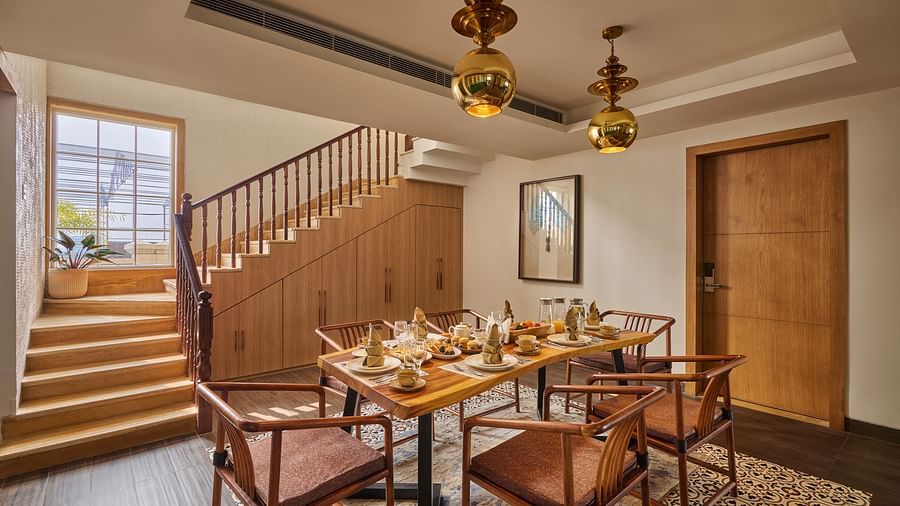 Modern dining room with a wooden table set for four, leather chairs, and stylish pendant lights. A staircase with wooden railings leads to an upper level, and natural light enters through a large window beside the stairs - Voyage The Solitaire Bliss.