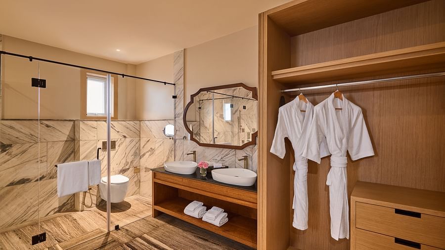 Modern bathroom with a marble-tiled shower area enclosed by glass, a double sink vanity with a wooden base, and a large decorative mirror. An open wooden wardrobe displays neatly hung white robes and folded towels - Voyage The Solitaire Bliss.
