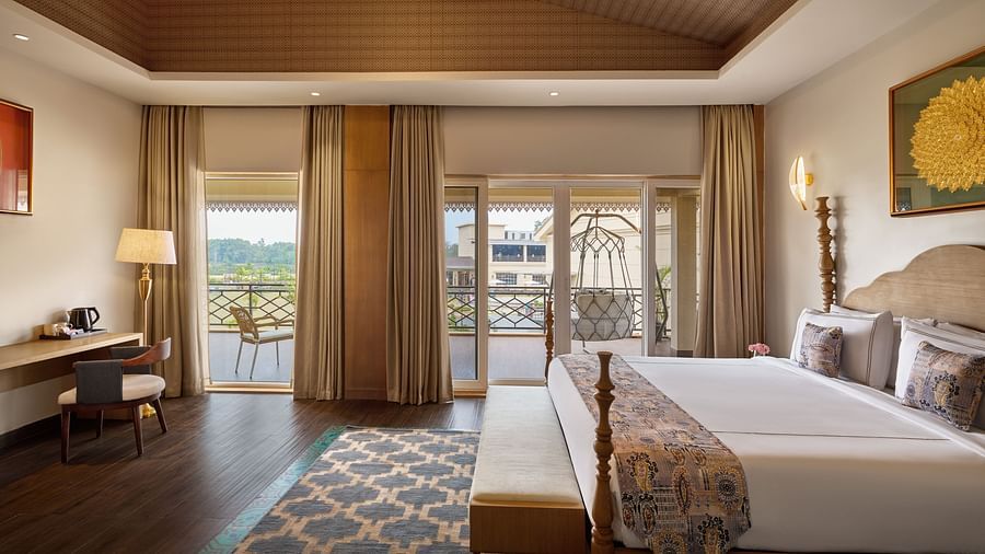 A well-lit hotel room featuring a king size bed with a patterned throw, a wooden desk and chair, and a balcony with a swing offering a view of a cityscape. The room has a wooden ceiling and a patterned rug - Voyage The Solitaire Bliss.