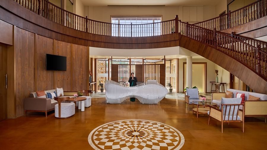 A spacious resort lobby features a high ceiling and wooden staircases curving on either side. A circular patterned tile lies on the polished floor in front of a white, wave-like reception desk where a person stands - Voyage The Solitaire Bliss.