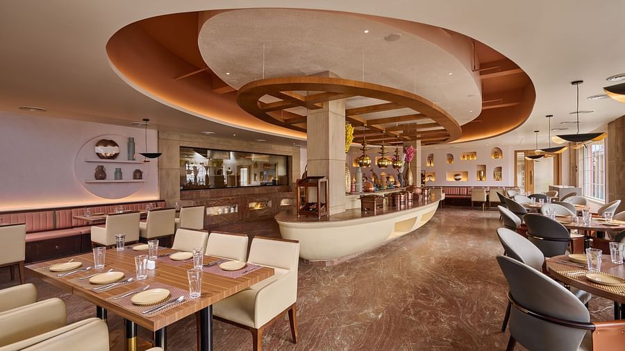 A stylish restaurant interior showcases a centrally located, elliptical bar with hanging pendant lights. Dining tables with set places are arranged throughout the space, which features a patterned marble floor and warm lighting - Voyage The Solitaire Bliss