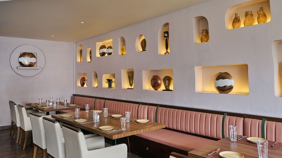 A restaurant dining area shows tables set with plates and glasses, positioned next to a long banquette with pink upholstery. The textured white wall is decorated with illuminated niches displaying various decorative objects - Voyage The Solitaire Bliss.