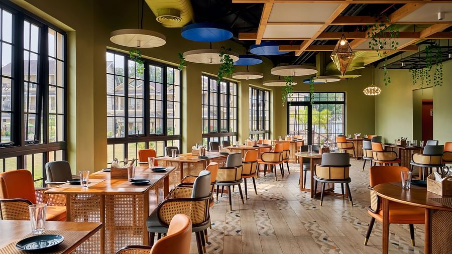 Vibrant and airy restaurant with stylish wooden furnishings, rattan-accented chairs, large windows offering natural light, and a modern ceiling with hanging greenery - Voyage The Solitaire Bliss.