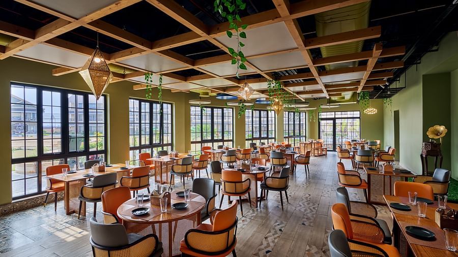 Stylish restaurant interior with round wooden tables, cushioned chairs, large grid windows, and a decorative wooden ceiling featuring hanging plants and pendant lights - Voyage The Solitaire Bliss.