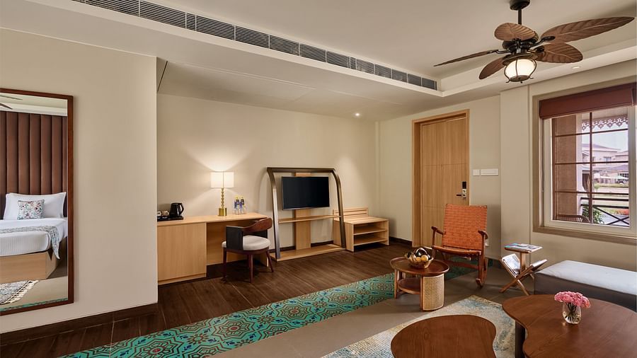 Spacious hotel room with modern wooden furnishings, a work desk with a TV, ceiling fan, cosy seating area, and a bed partially visible in the mirror reflection - Voyage The Solitaire Bliss.
