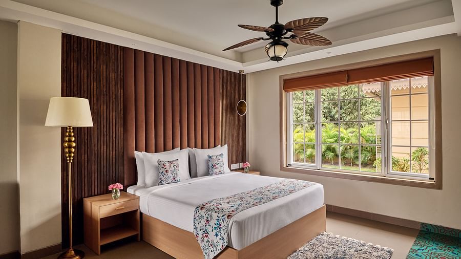 A bedroom with a king bed featuring white linens and a patterned throw, flanked by a wooden nightstand with a lamp and a textured brown headboard. A ceiling fan hangs above, and a large window with blinds reveals a view of lush greenery outside. A patterne