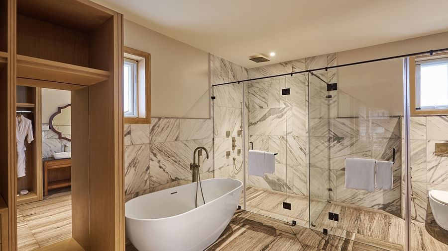 A spacious bathroom with marble-tiled walls features a freestanding white tub, a glass-enclosed shower with silver fixtures, and light wood flooring. Two windows fill the room with natural light, while open wooden shelves reveal a separate sink and mirror 