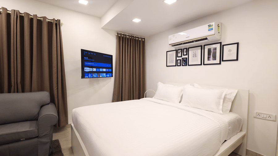 A bedroom at Wabi - Sabi by Alamiko, Juhu, featuring a plush bed, a sofa, framed painting on a wall, an A/C unit, and a smart TV.