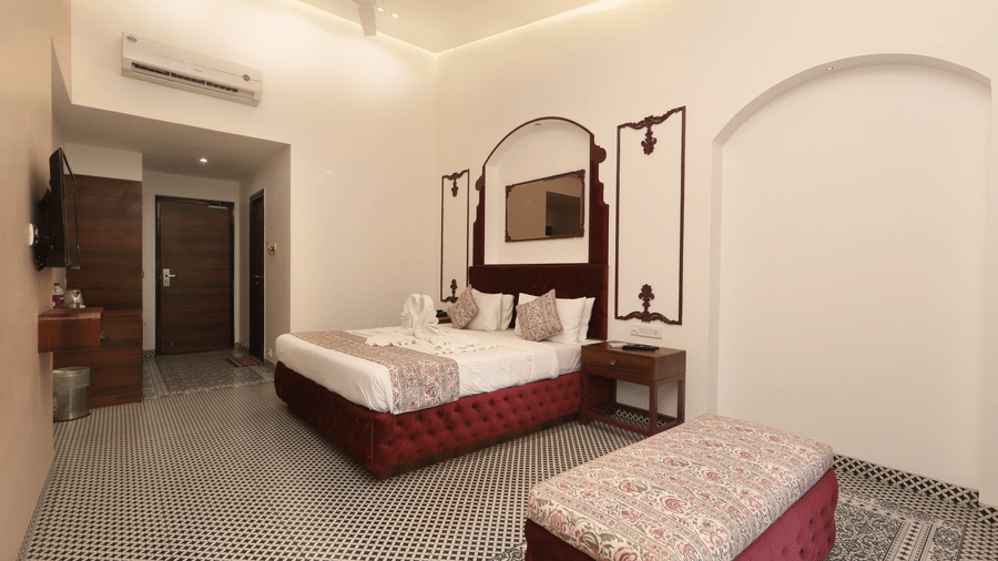 Elegant hotel bedroom with double bed, patterned bedding, bedside lamps, decorative mirror with arches, cushioned bench, tiled floor, and warm ambient lighting.