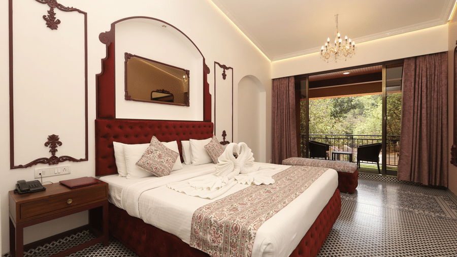 Moghul Room at Zaras Resort with elegant décor, red accents, swan towel art on bed, balcony view of greenery, and ambient lighting.