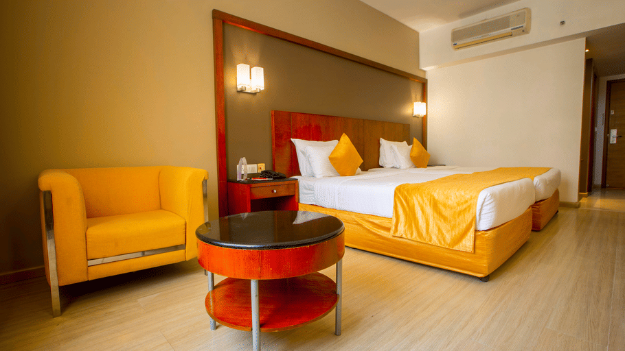 Hotel room at Featherlite Evoma Hotel with a bed dressed in yellow and white linen, a yellow armchair and a round red side table nearby.