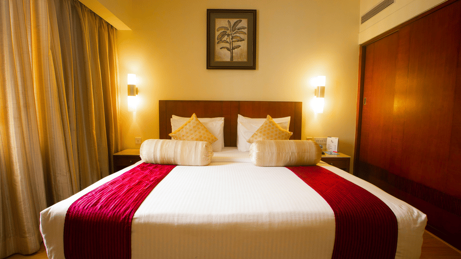 Hotel room at Featherlite Evoma Hotel with a bed dressed in white and maroon linen, a wooden headboard and framed wall art placed nearby.