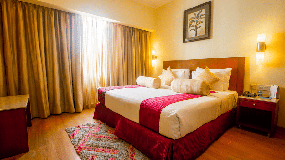 Hotel room at Featherlite Evoma Hotel with a bed dressed in white and maroon linen, wooden furnishings and long curtains by the window nearby.