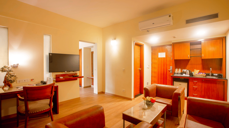 Suite living area at Featherlite Evoma Hotel with a red sofa, a wall-mounted television, a kitchenette and warm wooden flooring below.