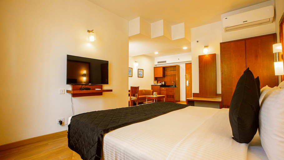 Hotel room at Featherlite Evoma Hotel with a bed dressed in white and dark grey linen, a wall-mounted television and a seating area nearby.