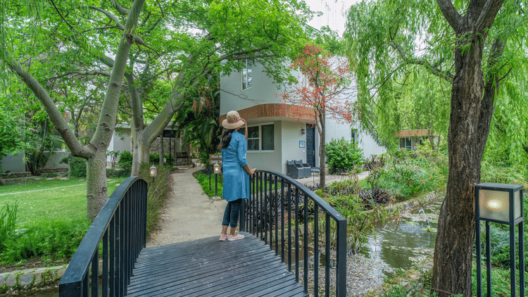 A bridge with a women standing on it which is over a small pond leading to a building surrounded by trees and greenery at The Golden Tusk, Jim Corbett