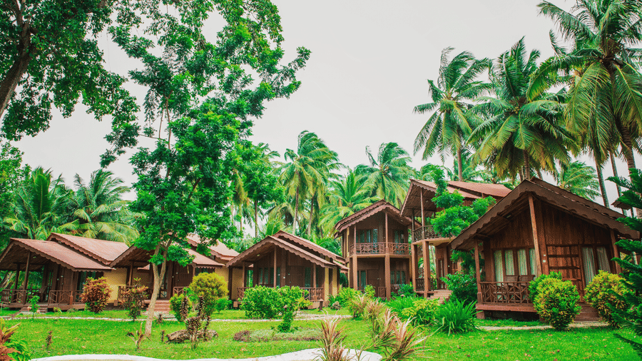 multiple rustic wooden cabins nestled in a lush, tropical landscape surrounded by tall green trees and palm trees under a pale sky in Silver Sand Beach Resort