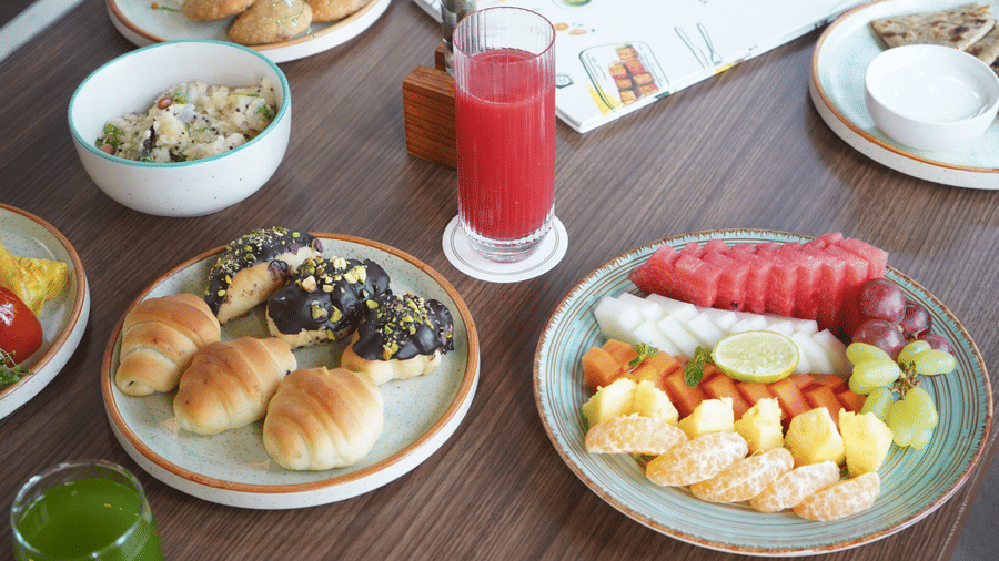 A dining table set with multiple plates of food, including a plate of small pastries, a plate of sliced fruit, and a glass of juice at The Golden Tusk, Jim Corbett