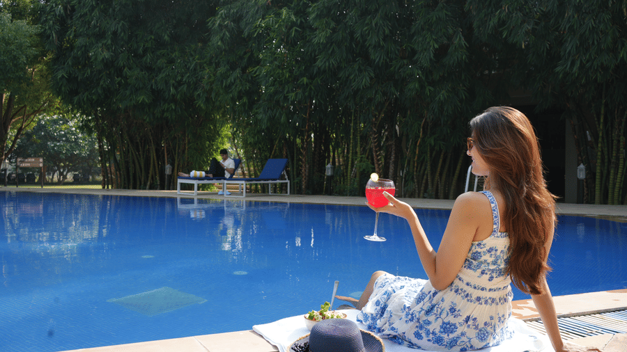 A woman in a floral dress sitting by the edge of a swimming pool with a drink during the day at The Golden Tusk, Jim Corbett