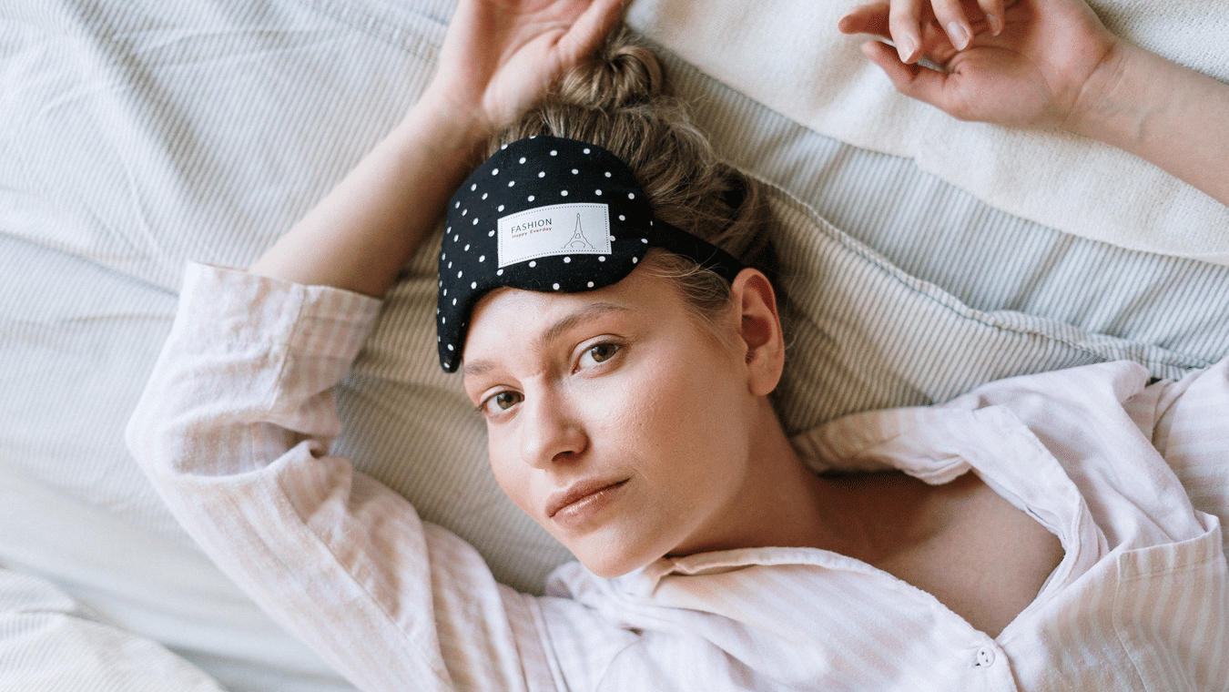 A woman lying in bed with her hands over her head and a sleeping mask on her forehead