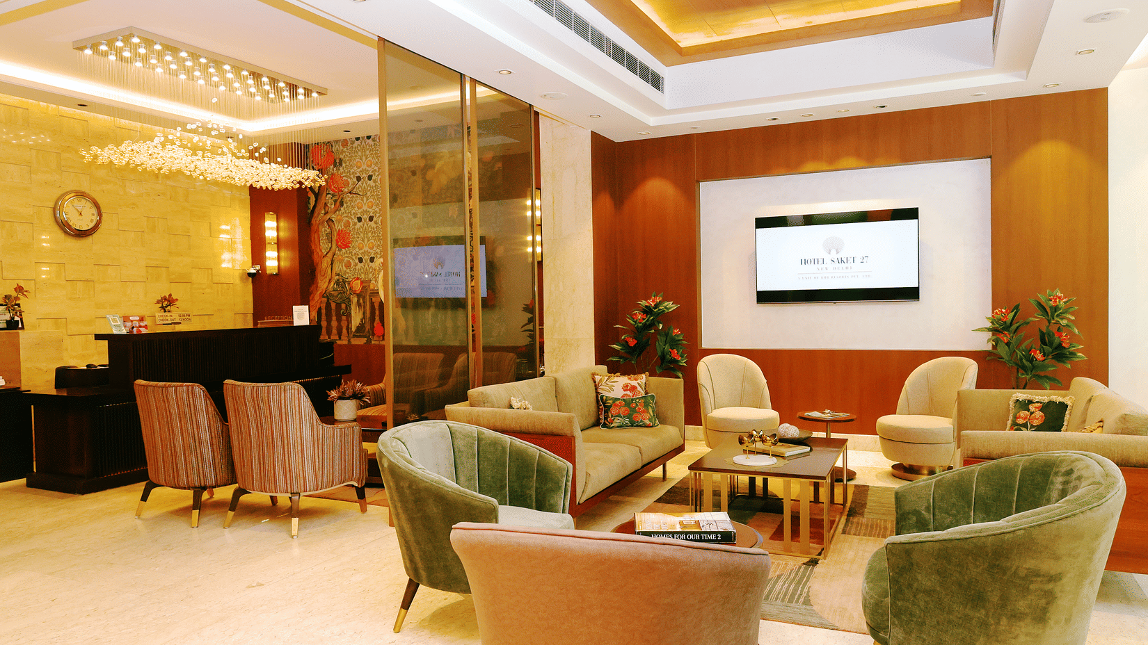 Lounge area at Hotel Saket 27 with reception desk, cushioned chairs, sofas, centre tables, wall mounted television, indoor plants and ceiling lighting.