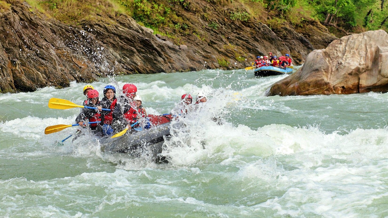 A group of people wearing red life jackets and helmets rafting down a fast-moving river surrounded by lush, green hills.