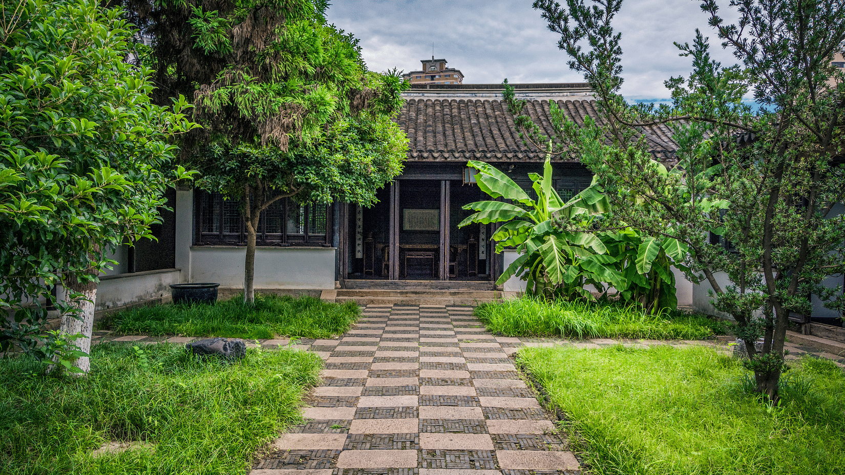 A traditional courtyard with a tiled-roof building, lush greenery, and a patterned stone pathway in the foreground.
