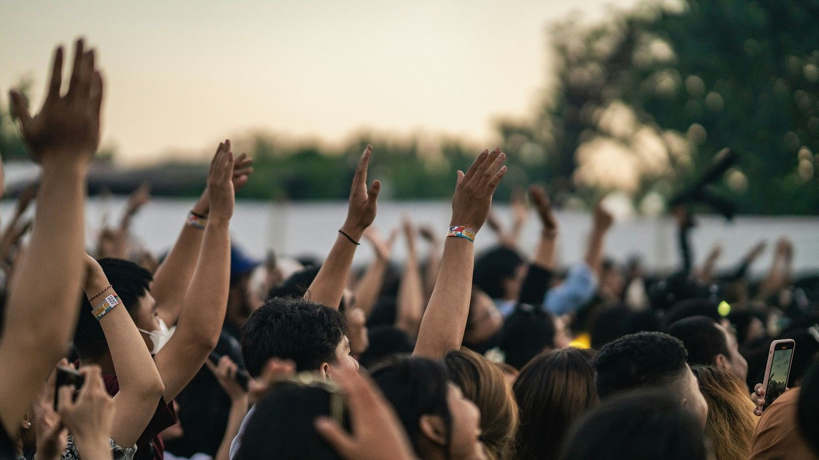 A lively crowd at an outdoor concert with raised hands, wristbands, and people capturing the moment on their phones.