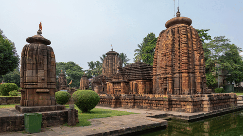 Ancient stone Mukteswar Temple, Bhubaneswar complex with carved towers, flags, and a water feature, surrounded by trees.